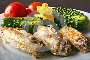 Baked poultry chicken wings, fresh natural tomatoes and green organic cucumbers on a white plate on a black background in closeup.