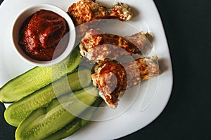 Baked poultry chicken wings, tomato paste and sliced cucumbers on a white plate on a black background. Top view from above. Copy s