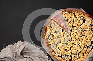 Baked round crumble pie with plum cut into pieces on a black background