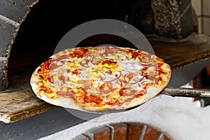 Baked salami pizza on metal paddle in front of wood oven.