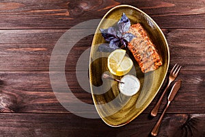 Baked salmon fillet with cheese sauce, basil and lemon on plate on wooden background. Hot fish dish.