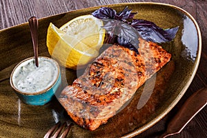 Baked salmon fillet with cheese sauce, basil and lemon on plate on wooden background. Hot fish dish. Top view.