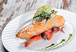 Baked salmon garnished with olives, greens, tomatoes on plate over wooden background. Hot fish dish