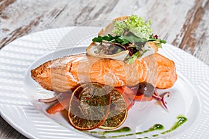 Baked salmon garnished with olives, greens, tomatoes on plate over wooden background. Hot fish dish