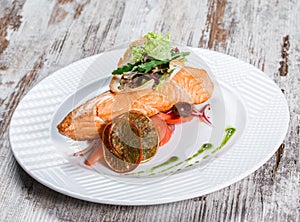 Baked salmon garnished with olives, greens, tomatoes on plate over wooden background. Hot fish dish