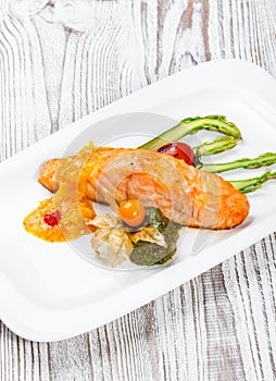 Baked salmon garnished with physalis, asparagus, tomatoes with herbs on wooden background.