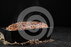 Baked wheat and rye bread with seeds in a baking dish, steam rises above a loaf, close-up with selective focus.