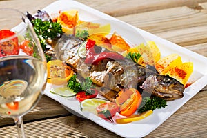 Baked whole trout with potatoes, greens and tomatoes on plate, glass of wine
