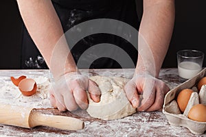 Baker in black apron prepares the dough on a wooden table, male hands knead the dough with flour