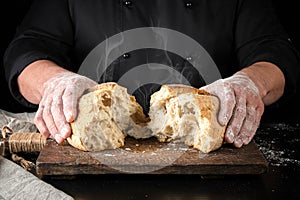 baker in black uniform broke in half a whole baked loaf of white wheat flour bread