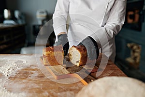 Baker chef holding knife and cutting fresh bread on wooden table