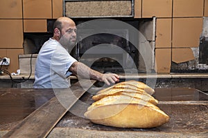 Baker. A young handsome bakery worker on the background of bread, takes bread from a stove with a wooden shovel. Industrial