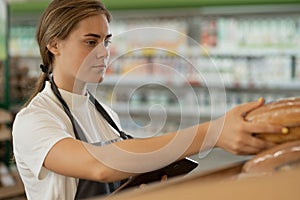 Bakery female worker in uniform putting loaf of bread on shelf in supermarket. In background shelf full with fresh