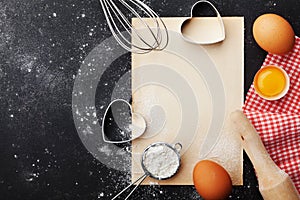 Baking background with flour, rolling pin, eggs, paper sheet and heart shape on kitchen black table from above for Valentines day.