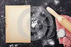 Baking background with flour, rolling pin, paper sheet and heart shape on kitchen black table from above for Valentines day.