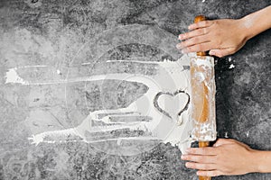 Baking background. Girl hands keep rolling pin and heart of flour on gray table with copy space, top view.