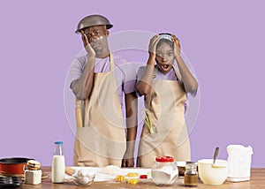Baking disaster concept. Desperate young black couple having difficulty cooking over lilac background