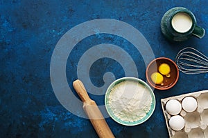 Baking Ingredient- flour, egg, milk, rolling pin, whisk on a blue painted background. Top view, flat lay, copy space