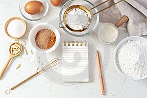 Baking pastry or cake ingredients with notebook and pen, sifting flour, brown sugar, eggs and milk with utensil on marble table