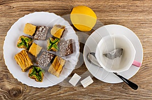 Baklava in white plate, lemon, sugar, cup with tea bag, spoon on saucer on table. Top view