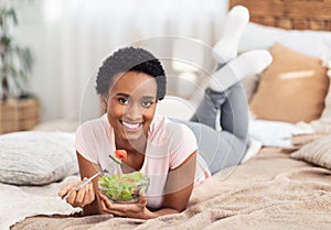Balanced raw food diet concept. Beautiful black woman eating fresh vegetable salad on bed at home, free space