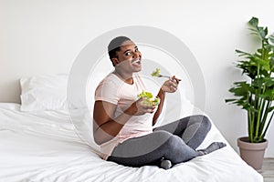 Balanced slimming diet concept. Overweight black woman eating vegetable salad on bed at home, full length