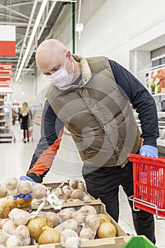 Bald adult male in medical mask and gloves chooses vegetables in a supermarket. Self-isolation regime during the coronavirus