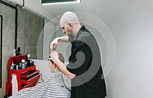 Bald barber clipping young client with scissors on white wall background. Professional male hairdresser creates a stylish