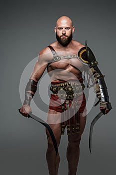 Bald and bearded gladiator poses standing tall with two swords