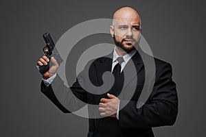 Bald and bearded man in a black suit holds a pistol, exuding power