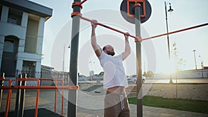 Bald and bearded man doing pull-ups on horizontal bar. On workout area near house