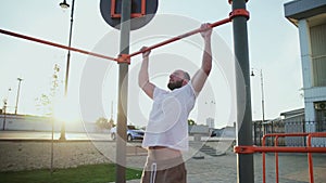 Bald and bearded man doing pull-ups on horizontal bar. On workout area near house