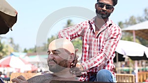 Bald bearded man get head, neck and body massage from worker in glasses at sunny beach in shadow, surfing net in phone