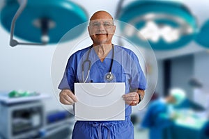 Bald caucasian male doctor wearing blue scrubs in an operating room holding blank white sign and smiling