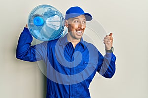 Bald courier man with beard holding a gallon bottle of water for delivery pointing thumb up to the side smiling happy with open