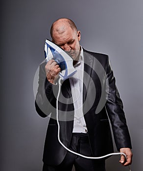 Bald fun tired unhappy loser failed business man punishing himself holding the home iron near the face in suit on grey background