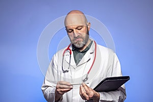 Bald hairless doctor on blue background. Male in his 40s with grey beard. Holding black file and looking at thermometer. Dramatic