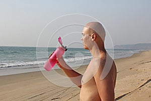 A bald, handsome man with a bare or bare torso is drinking water from a pink fitness bottle. The man is thirsty. Heat and thirst.