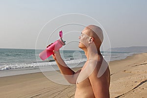 A bald, handsome man with a bare or bare torso is drinking water from a pink fitness bottle. The man is thirsty. Heat and thirst.