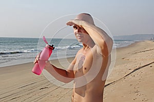 A bald, handsome man with a bare or bare torso is drinking water from a pink fitness bottle. The man is thirsty. Heat and thirst.