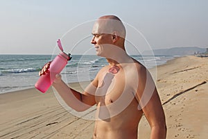 A bald, handsome man with a bare or bare torso is drinking water from a pink fitness bottle. The man is thirsty. Heat and thirst.