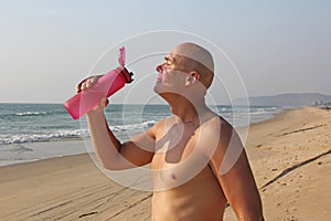 A bald, handsome man with a bare or bare torso is drinking water from a pink fitness bottle. The man is thirsty. Heat and thirst.