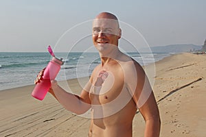 A bald, handsome man with a bare or bare torso is drinking water from a pink fitness bottle. The man is thirsty. Heat and thirst.