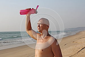 A bald, handsome man with a naked or bare torso watering himself with water from a pink fitness bottle. The man is thirsty. Heat