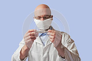 Bald male doctor looking at thermometer. Man wearing mask and glasses, light blue background. White uniform and blue shirt