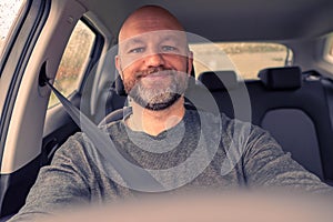 Bald male driver in focus, steering wheel out of focus. Man wears grey shirt, unshaven with grey and black beard in his 40s. Toned