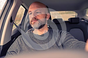 Bald male driver in focus, steering wheel out of focus. Man wears grey shirt, unshaven with grey and black beard in his 40s. Toned