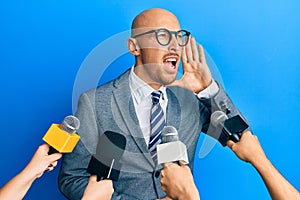 Bald man with beard being interviewed by reporters holding microphones shouting and screaming loud to side with hand on mouth