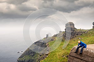Bald man with beard in blue jacket sitting on an edge of a cliff and looking down, Cliff of Moher, county Clare, Ireland, Low
