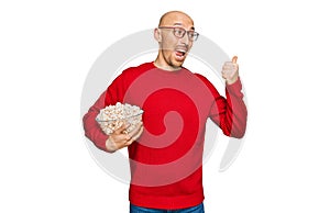 Bald man with beard eating popcorn pointing thumb up to the side smiling happy with open mouth